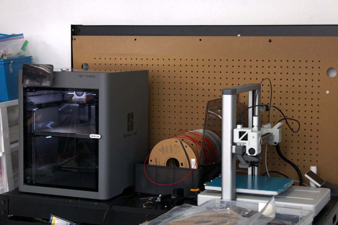 The workshop bench: a Bambu Lab P2S printer beside a smaller
                  printer, with spools of filament and a pegboard behind them